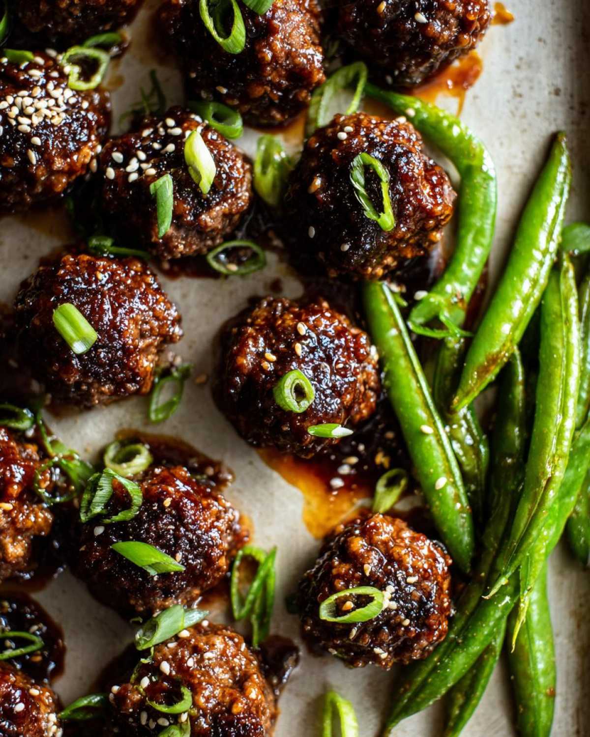 Close-up of Sheet Pan Sticky Sesame Meatballs coated in glaze, garnished with sesame seeds and green onions, next to bright green snap peas.