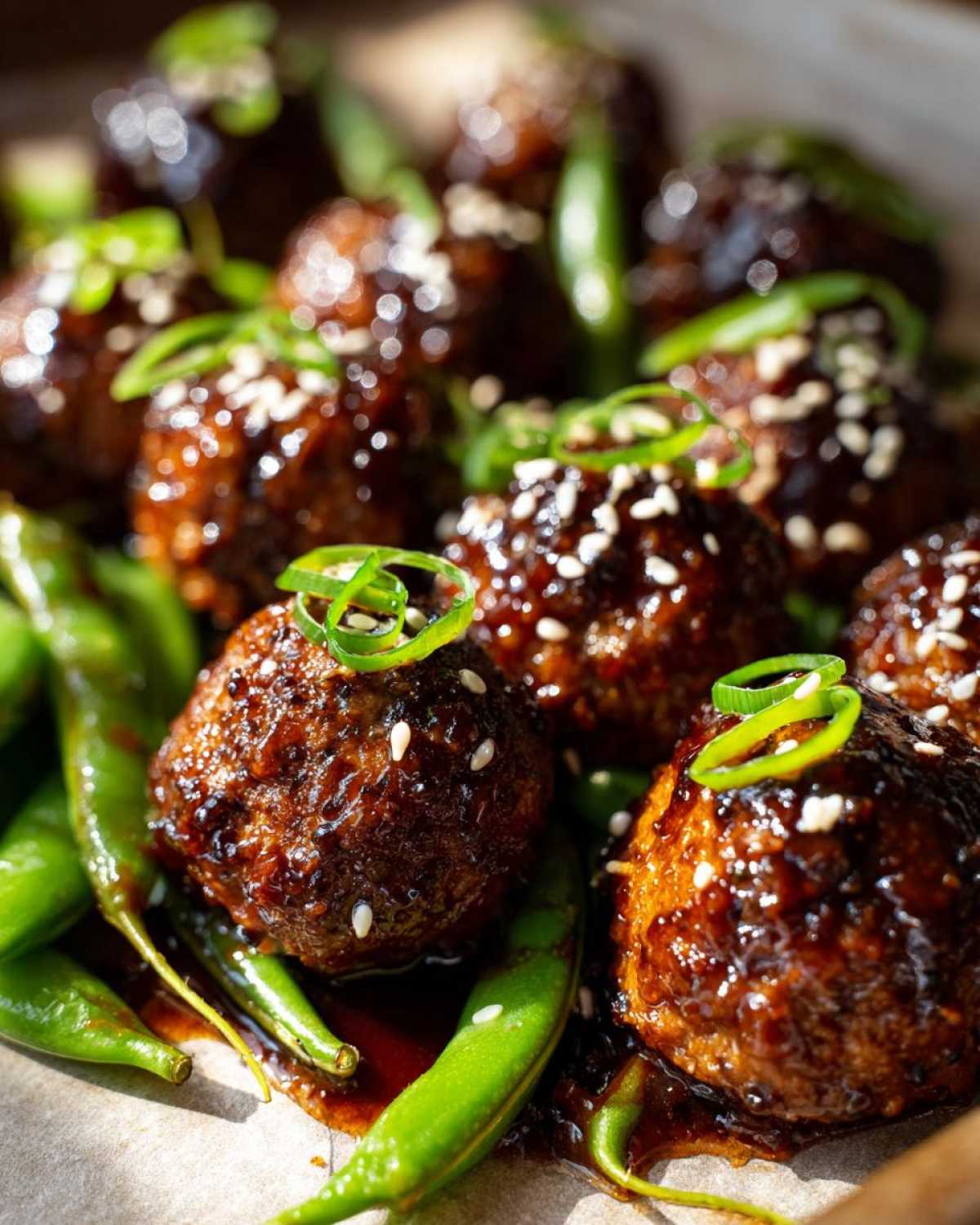 Close-up of Sheet Pan Sticky Sesame Meatballs coated in glaze, garnished with sesame seeds and green onions, served over bright green snap peas.