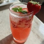 A tall glass filled with vibrant pink Sparkling Strawberry Basil Lemonade, garnished with fresh basil leaves and a whole strawberry.
