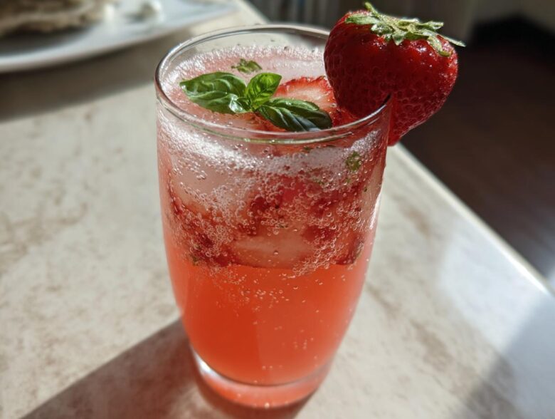 A tall glass filled with vibrant pink Sparkling Strawberry Basil Lemonade, garnished with fresh basil leaves and a whole strawberry.