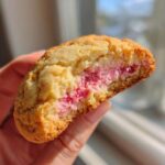 A hand holding a halved Strawberry Cheesecake Cookies showing the soft, golden exterior and bright pink, creamy strawberry filling.