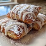 A stack of three golden-brown Vanilla French Beignets heavily dusted with powdered sugar and drizzled with vanilla glaze.