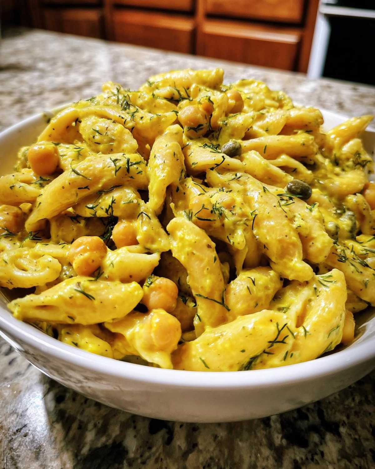 Close-up of Vegan Creamy Lemon Chickpea Pasta Salad in a white bowl, coated in yellow sauce and topped with dill.