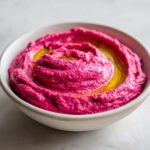 Close-up of bright pink Vegan Creamy Roasted Beet Hummus swirled in a white bowl, topped with olive oil.