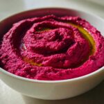 Close-up of vibrant magenta Vegan Creamy Roasted Beet Hummus swirled in a white bowl and drizzled with olive oil.