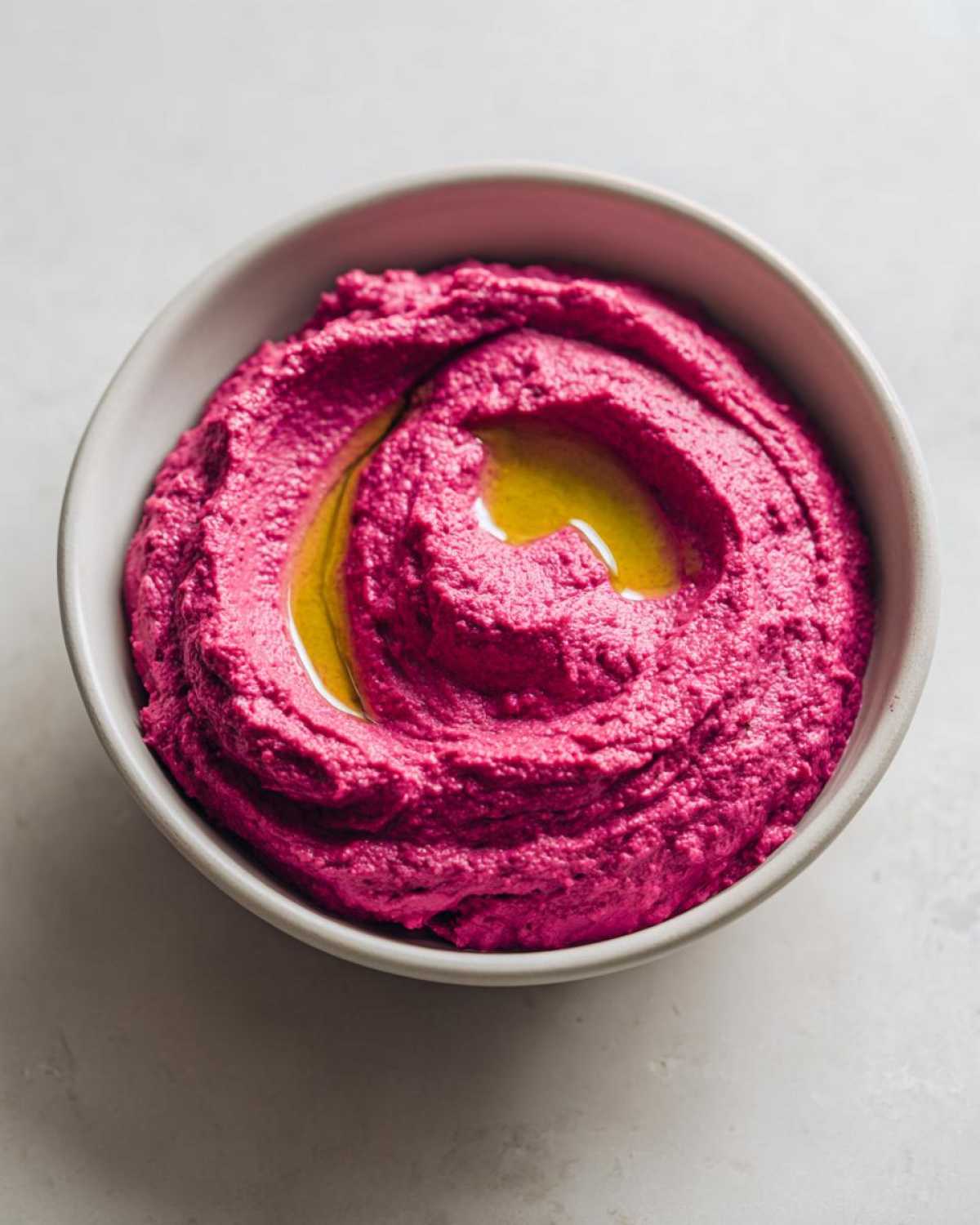 A close-up overhead shot of vibrant pink Vegan Creamy Roasted Beet Hummus swirled in a grey bowl, drizzled with olive oil.