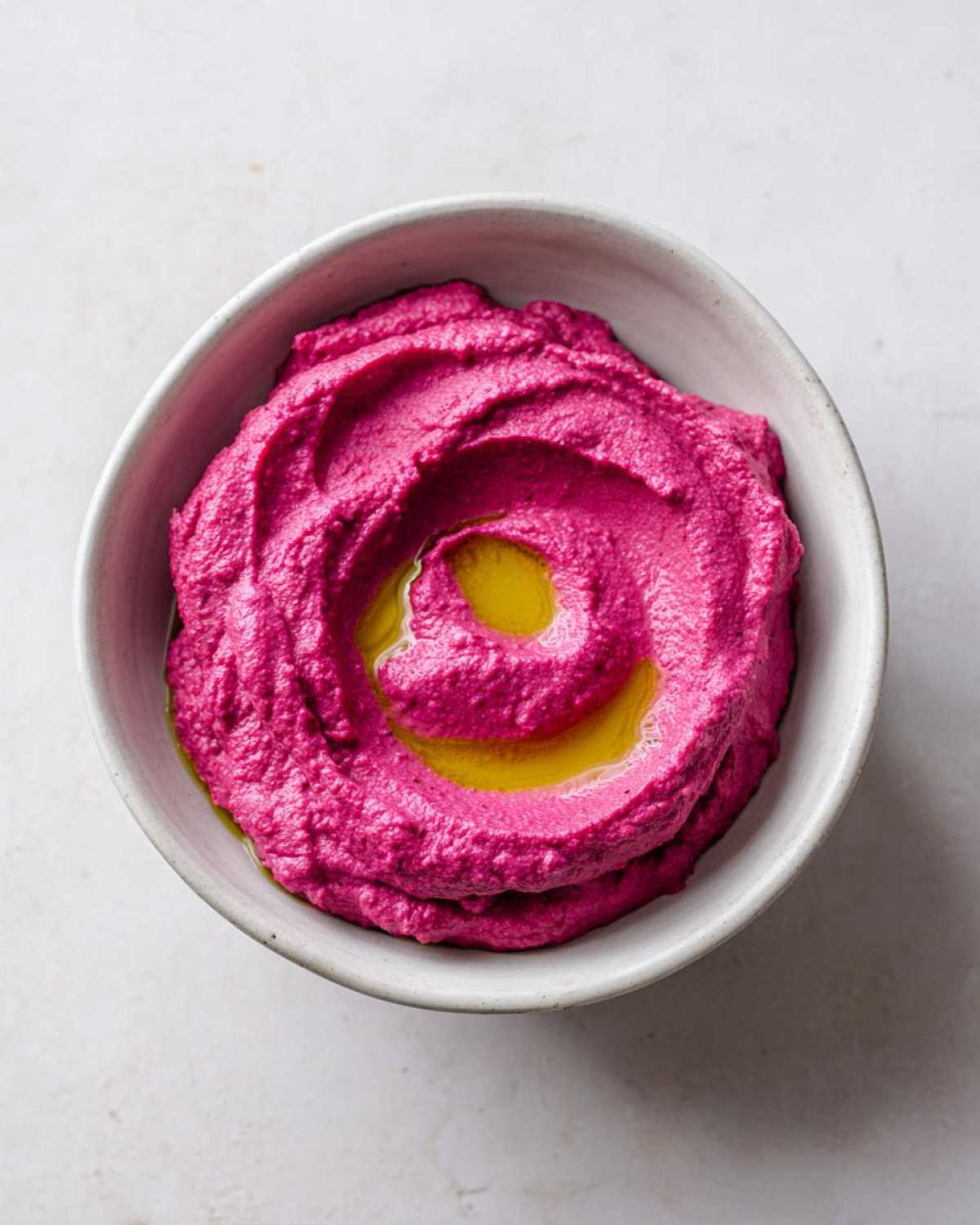 Overhead shot of bright pink Vegan Creamy Roasted Beet Hummus swirled in a light bowl, drizzled with olive oil.