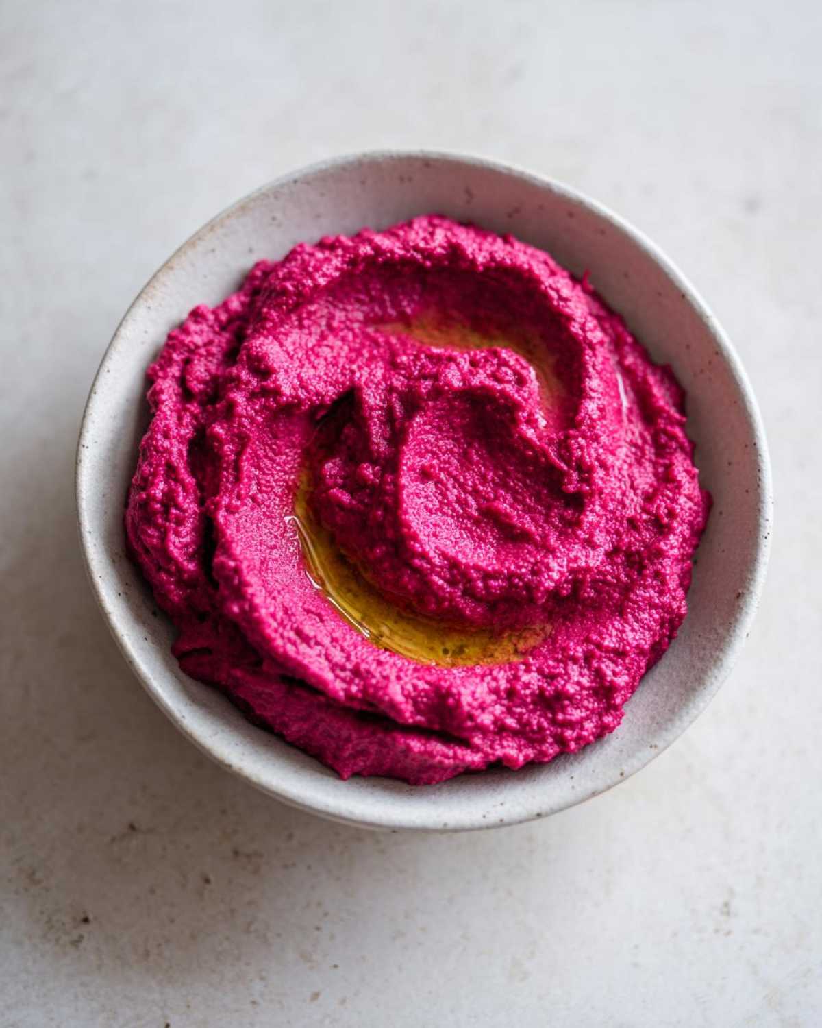 A close-up overhead shot of bright magenta Vegan Creamy Roasted Beet Hummus swirled in a rustic bowl, drizzled with olive oil.