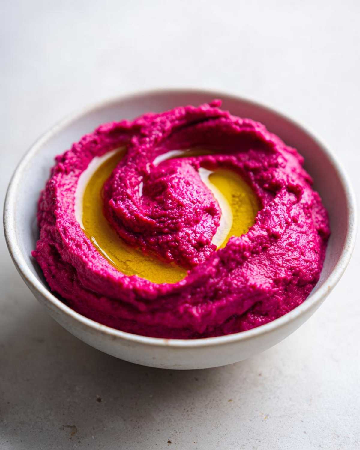 Close-up of vibrant magenta Vegan Creamy Roasted Beet Hummus swirled with olive oil in a light ceramic bowl.