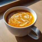 Close-up of vibrant orange Vegan Creamy Roasted Carrot Ginger Soup topped with a swirl of white vegan cream.