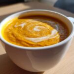 Close-up of a vibrant orange Vegan Creamy Roasted Carrot Ginger Soup topped with a swirl of white cream.
