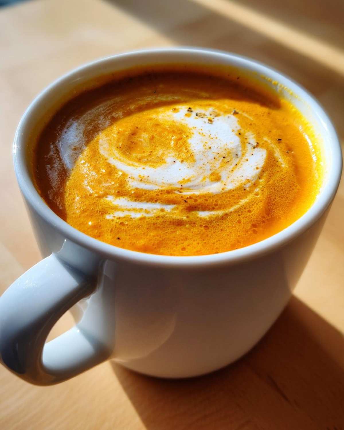 Close-up of bright orange Vegan Creamy Roasted Carrot Ginger Soup topped with a swirl of white cream and pepper.
