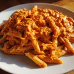 A close-up of penne pasta coated in a rich, orange Vegan Creamy Roasted Red Pepper Hummus Pasta sauce on a white plate.