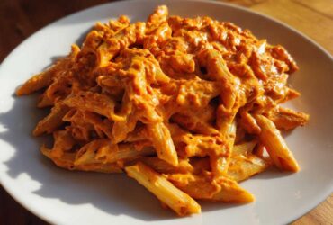 A close-up of penne pasta coated in a rich, orange Vegan Creamy Roasted Red Pepper Hummus Pasta sauce on a white plate.