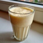 A close-up of a creamy, frothy Banana Chamomile Tea Smoothie served in a clear, ribbed glass.
