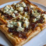 Two golden, flaky Caramelized Onion and Goat Cheese Tarts topped with crumbled cheese and fresh thyme sprigs.