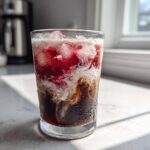 A close-up of a Cherry Vanilla Iced Latte showing dark coffee, white cream swirl, and bright red cherry syrup mixing over ice.