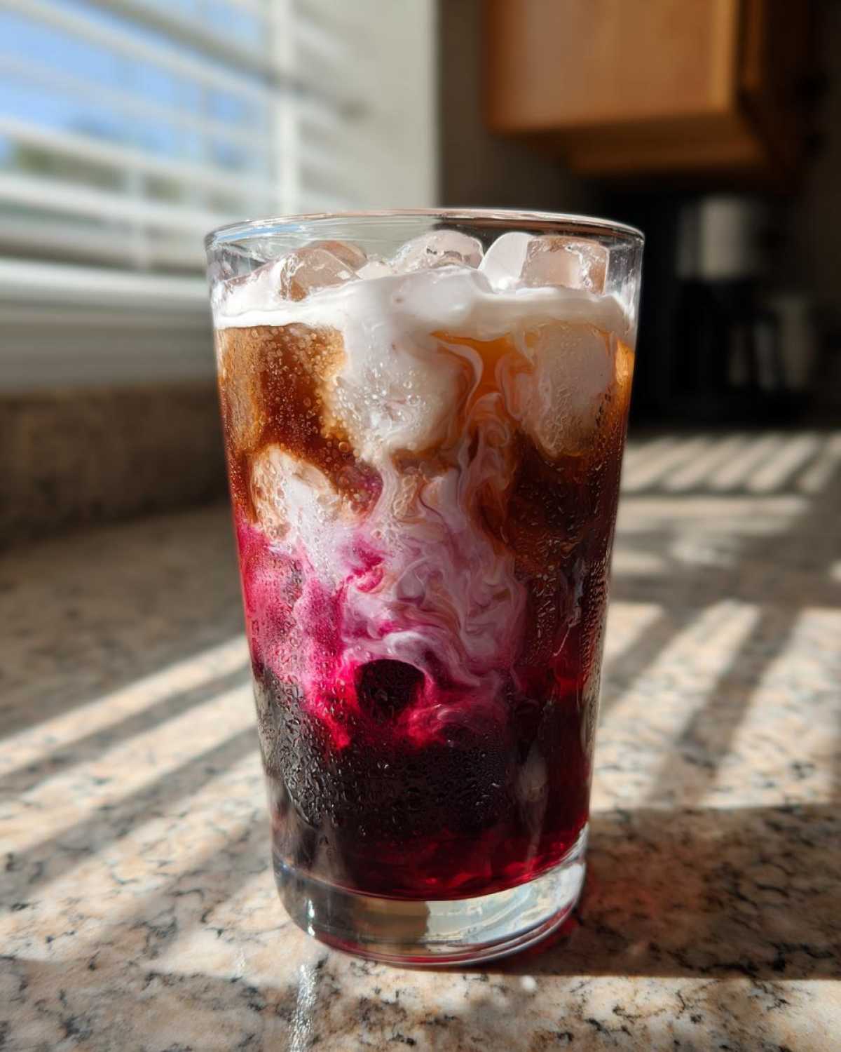 A tall glass showing the beautiful layers of a Cherry Vanilla Iced Latte with dark coffee, bright pink cherry syrup, and swirling vanilla cream.