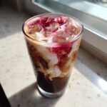 A tall glass of Cherry Vanilla Iced Latte showing layers of dark coffee, swirling cream, and bright red cherry syrup over ice.