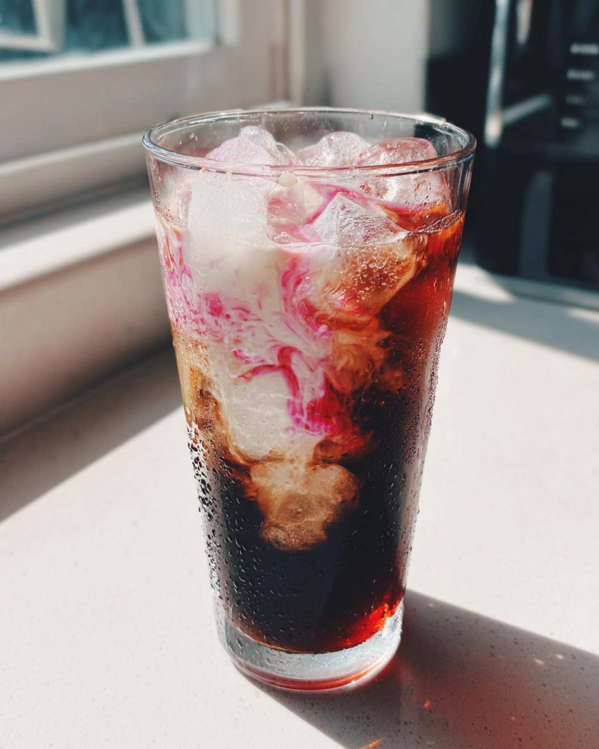 A tall glass of Cherry Vanilla Iced Latte with ice, showing creamy white milk swirling with bright pink cherry syrup.