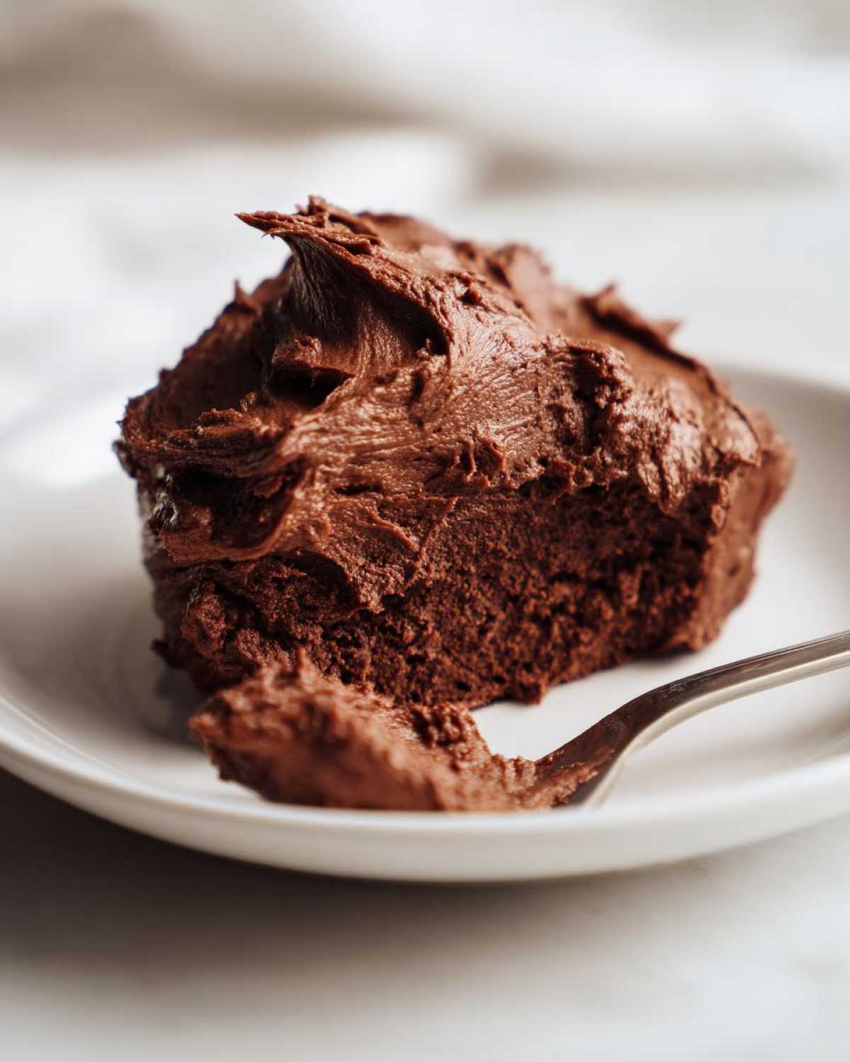 Close-up of a bite of cake generously topped with thick, rich Chocolate Buttercream Frosting on a white plate.