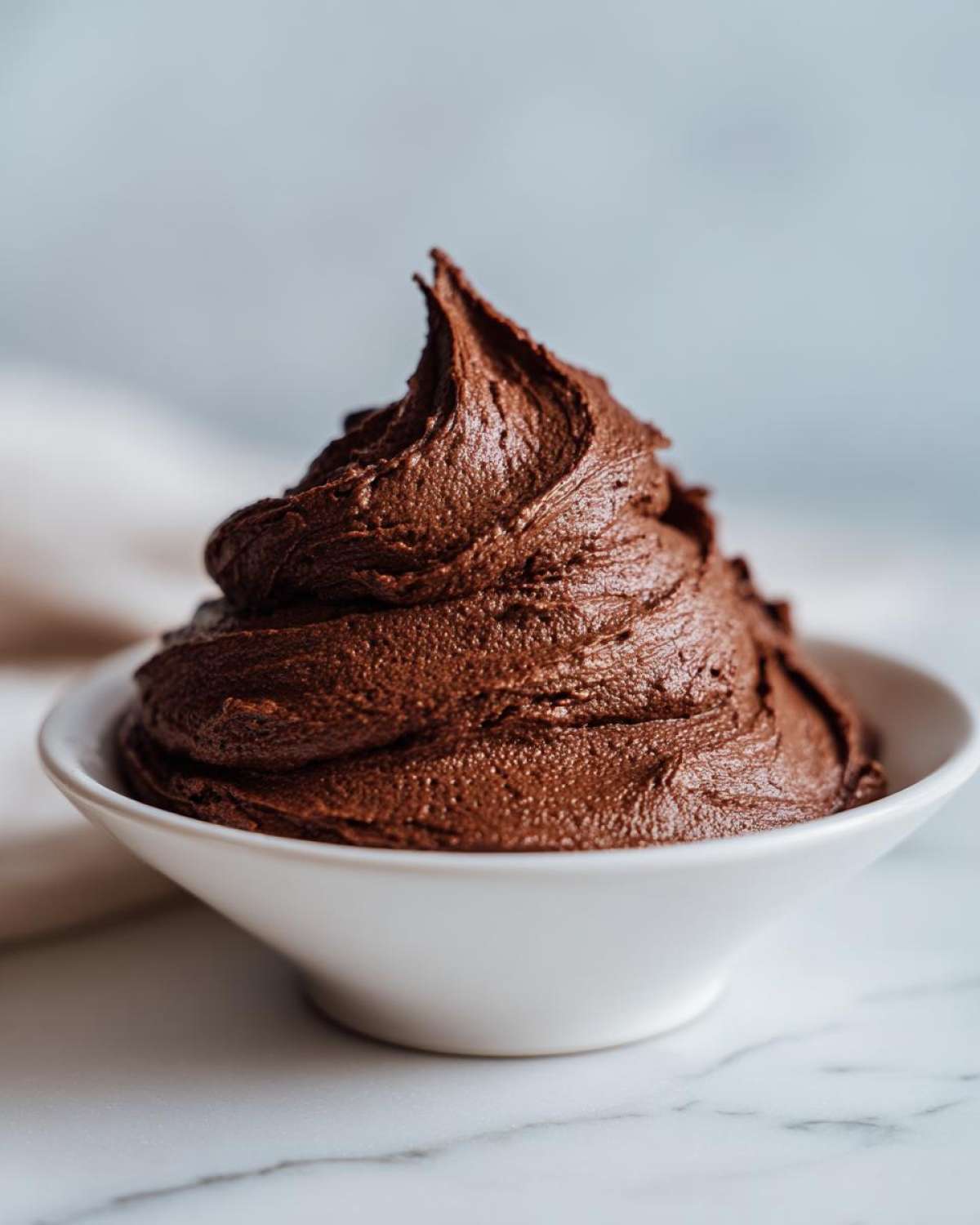A beautiful swirl of rich, dark Chocolate Buttercream Frosting piled high in a small white bowl.