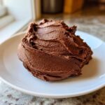 A close-up, appetizing swirl of rich Chocolate Buttercream Frosting resting on a small white plate.