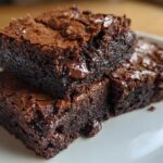 Close-up of two stacked, fudgy brownies with shiny, crackly tops and visible chocolate chips.