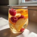 Close-up of a glass filled with bubbly White Peach Sangria, featuring raspberries, peach slices, and orange segments.