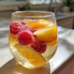Close-up of a glass filled with bubbling White Peach Sangria, featuring raspberries, orange slices, and peach pieces.