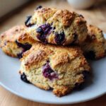 A stack of four freshly baked, golden-brown Blueberry Scones bursting with juicy blueberries, served on a light speckled plate.