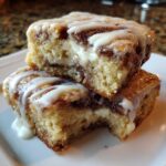 Two stacked squares of gooey Cinnamon Roll Bliss Bars with cream cheese filling and white icing drizzle.