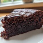 Close-up of a moist slice of Gluten Free Chocolate Cake with a thin, shiny chocolate glaze on top, served on a white plate.