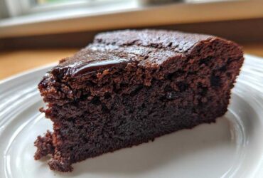 Close-up of a moist slice of Gluten Free Chocolate Cake with a thin, shiny chocolate glaze on top, served on a white plate.