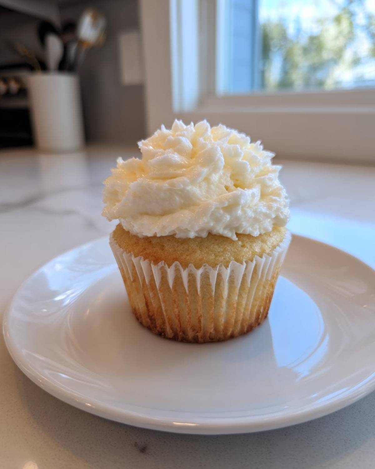 A single vanilla cupcake with a generous swirl of white frosting sitting on a small white plate.