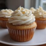 A close-up of a perfect vanilla cupcake with tall, swirled cream frosting, sitting on a white plate with more cupcakes blurred in the background.