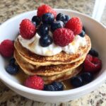 A stack of three golden Protein Pancake Bowls with Yogurt and Berries, topped with yogurt and fresh raspberries and blueberries.