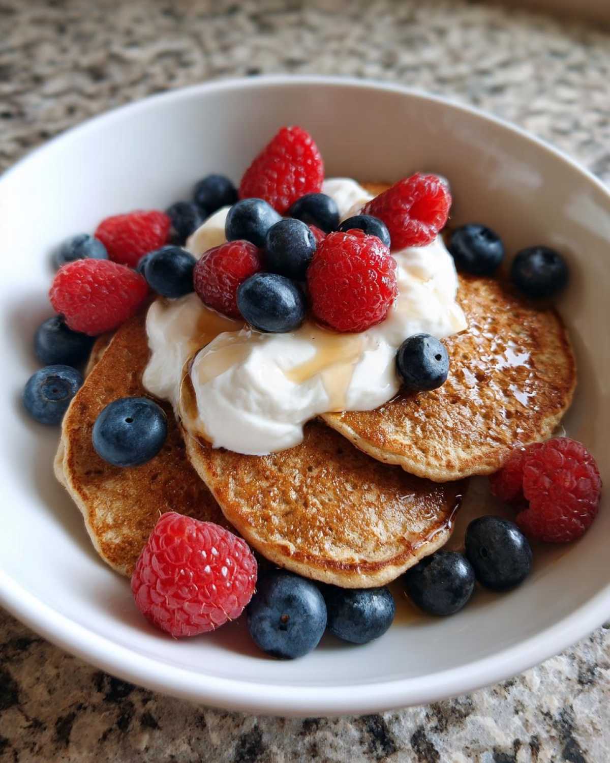 Close-up of Protein Pancake Bowls topped with yogurt, fresh raspberries, blueberries, and a drizzle of syrup.