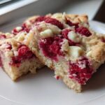Two moist squares of Raspberry White Chocolate Blondies, studded with bright red raspberries and chunks of white chocolate, served on a white plate.