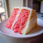 A close-up of a moist slice of pink Strawberry Cake with Cream Cheese Frosting on a white plate.